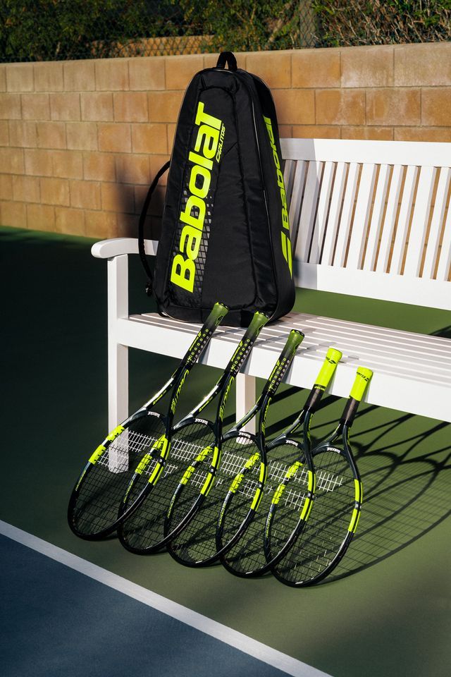 Schwarze Babolat-Tennistasche auf weißer Bank, davor fünf schwarz-gelbe Tennisschläger am Rand eines Hardcourts