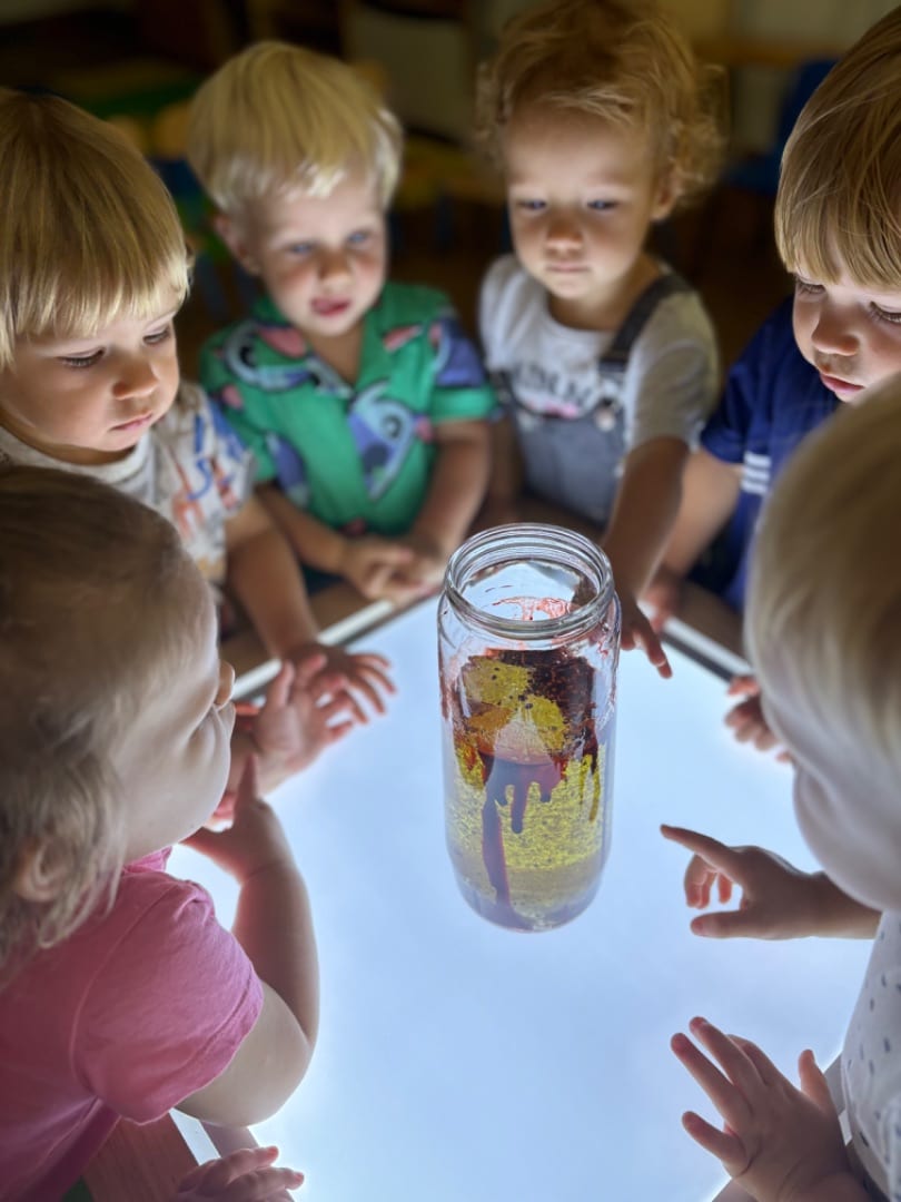Sześcioro dzieci stoi wokół podświetlonego stoika, na którym stoi słoik z kolorową masą.
