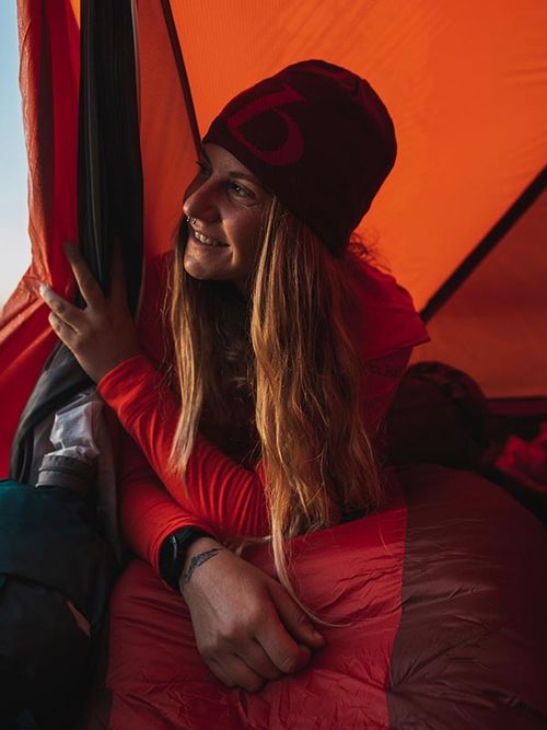 A person in a red beanie and jacket smiles while looking out from an orange tent, with a mountainous landscape in the background.