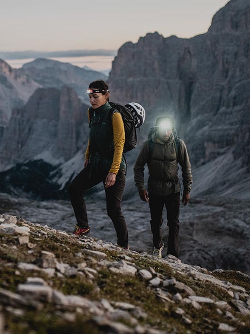 Two hikers ascend a rocky mountain trail at dusk, one with a headlamp illuminating the path, surrounded by towering cliffs.