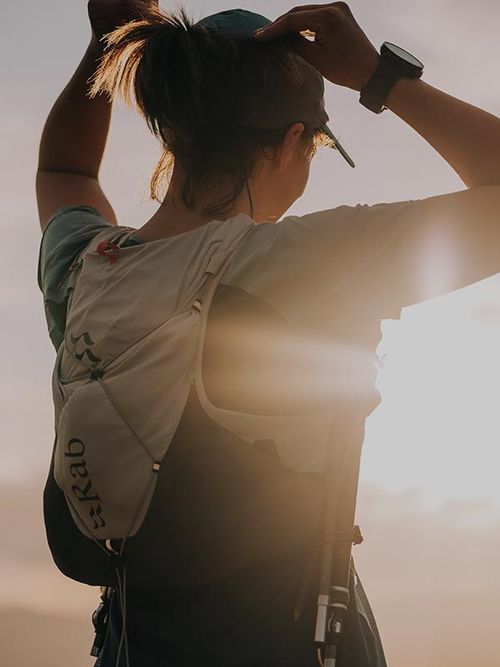 A person wearing a cap and running backpack facing the sunrise, with the sun creating a warm glow in the background.