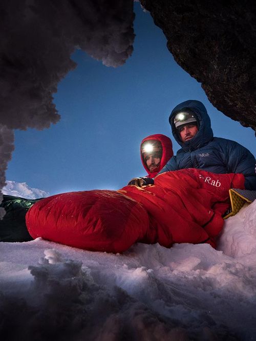 Two climbers in winter gear rest inside a snowy cave, illuminated by headlamps, with a red sleeping bag on the snow.