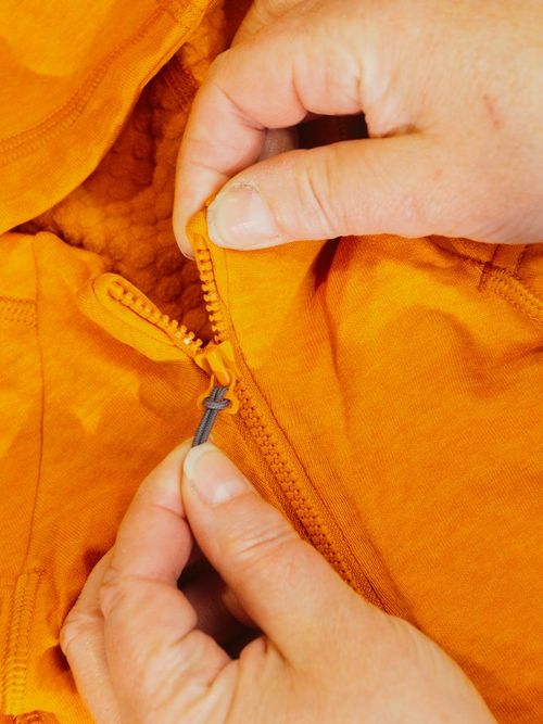 Hands zipping an orange jacket with a hood, showing the zipper detail and a small embroidered logo on the fabric.