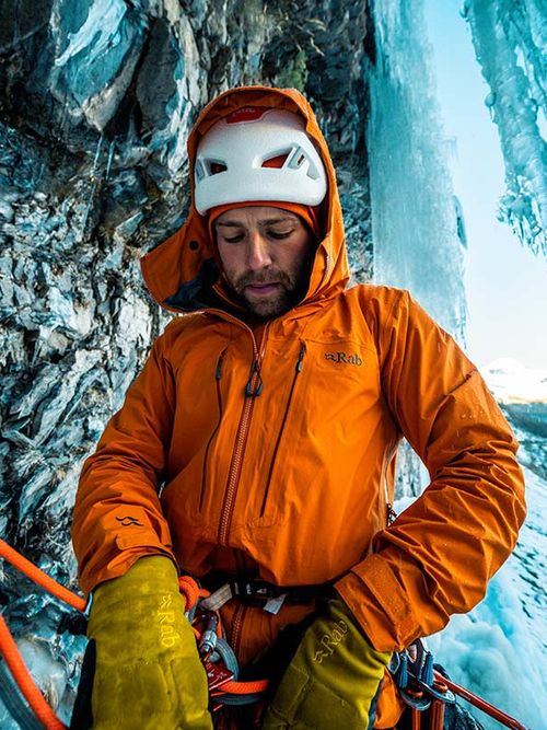 A climber in an orange jacket and helmet secures ropes on an icy rock face, surrounded by icicles and a clear blue sky.