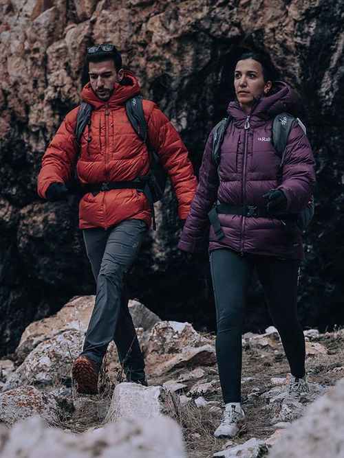 Two people hiking on rocky terrain, wearing backpacks and insulated jackets, one red and one purple, with a rocky background.