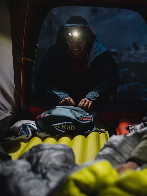 Person wearing a headlamp, smiling while packing a backpack inside a tent at night, with a sleeping bag and mat visible.
