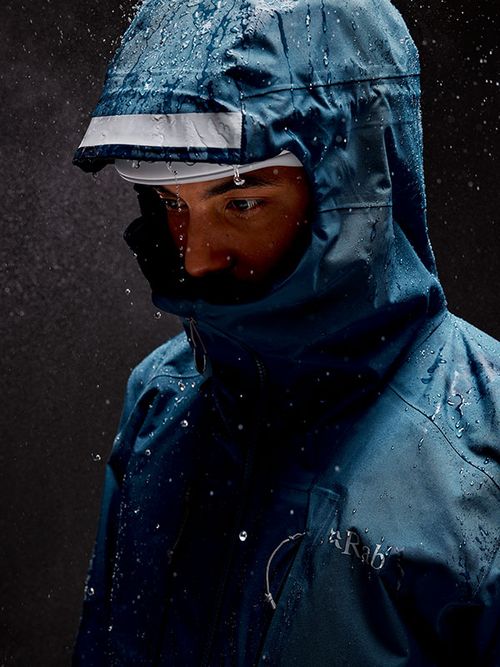 Person wearing a blue waterproof jacket with hood up, raindrops visible on the fabric, in a dark setting.