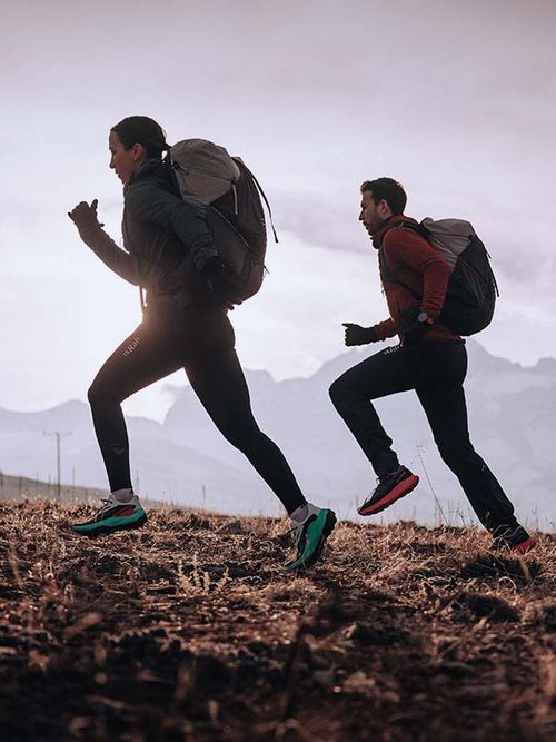 Two people with backpacks running uphill on a grassy trail, with misty mountains in the background.