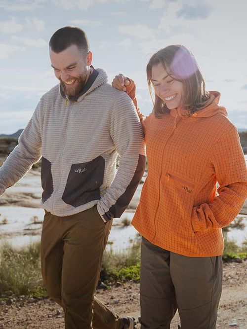 Two people walking outdoors, smiling, wearing hooded midlayers—one gray, one orange—against a bright, sunny backdrop.