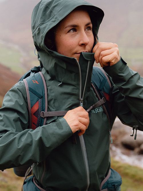 Person in a green rain jacket zipping up, wearing a backpack, standing outdoors with a misty, mountainous background.
