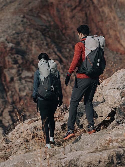 Two hikers with backpacks walking on a rocky trail, surrounded by rugged terrain and distant cliffs.