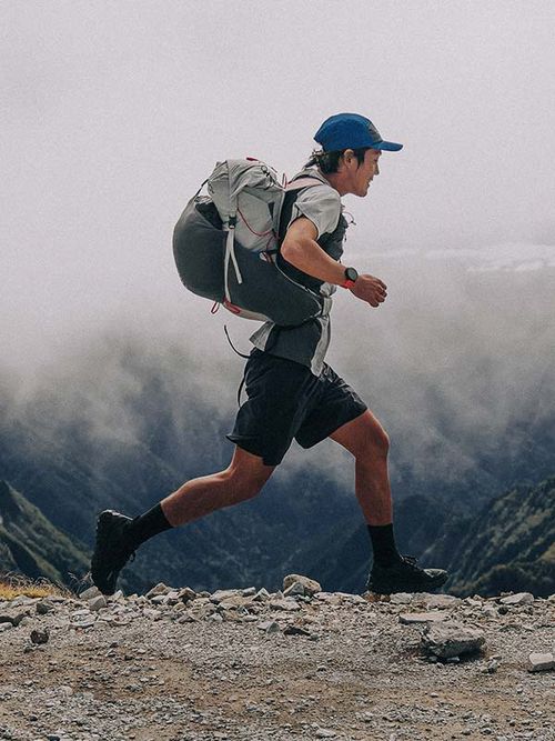 A person in hiking gear and a blue cap runs along a rocky mountain trail, with misty mountains in the background.