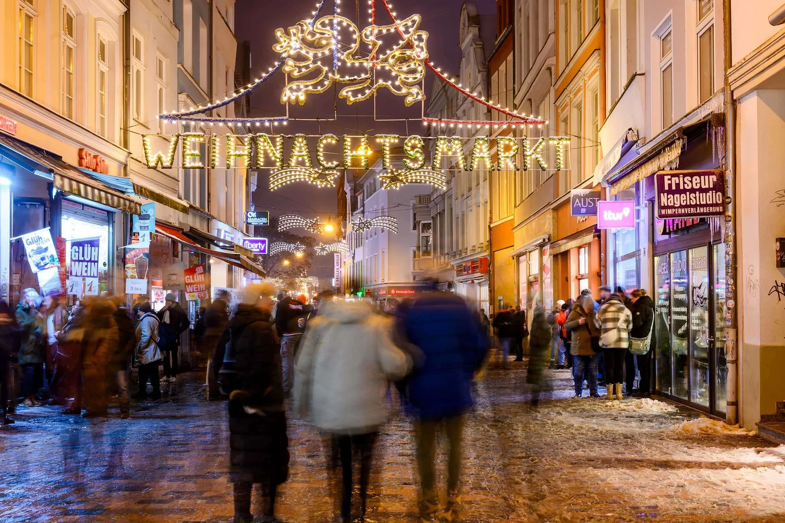 Schneebedeckte Straßen mit Weihnachtsmarktständen in Rostock, die festlich beleuchtet sind und eine warme, einladende Atmosphäre bieten – www.the-green.de.