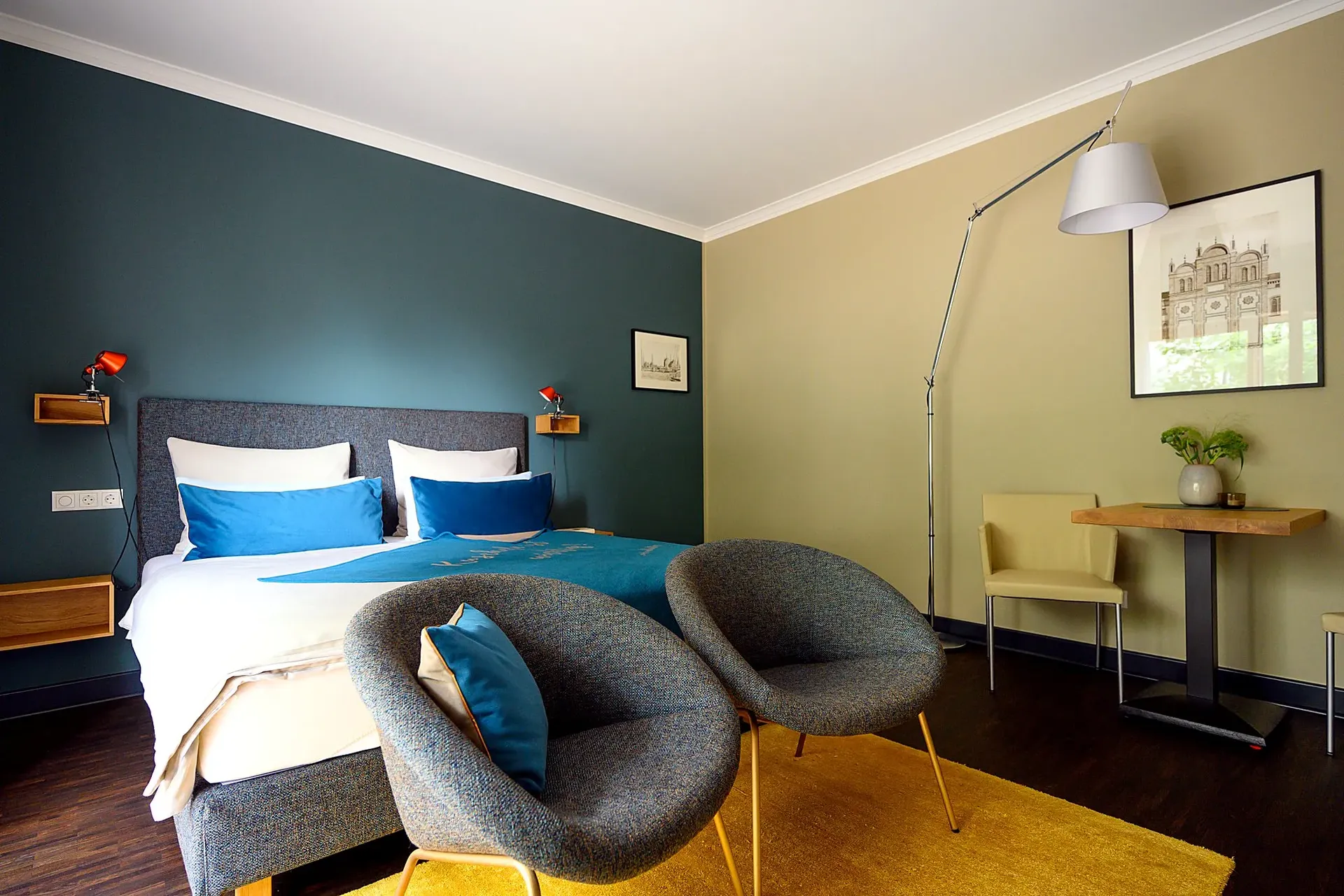 Schlafbereich mit petrolfarbener Wand, Doppelbett, zwei grauen Sesseln und Schreibtisch