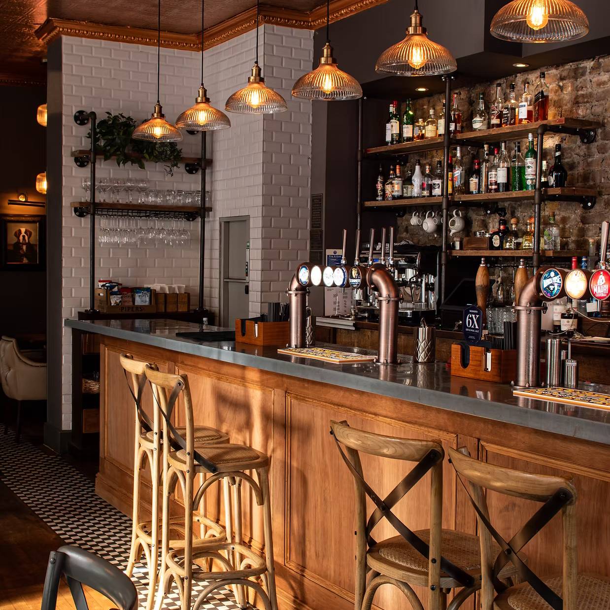 A cozy, well-lit bar interior with wooden stools, pendant lights, and a stocked back shelf. The checkered floor and brick wall add a rustic charm.