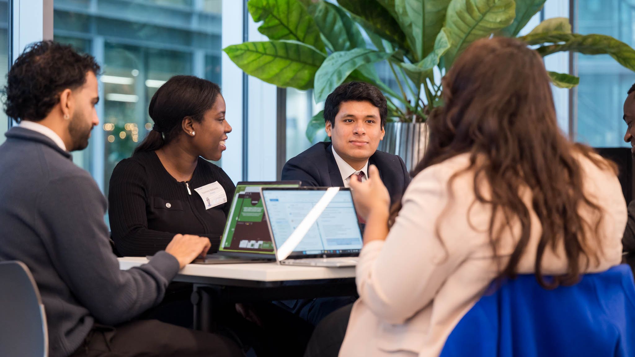 A group of people in a modern office setting engage in a discussion around a table, with laptops open and a large plant in the background.