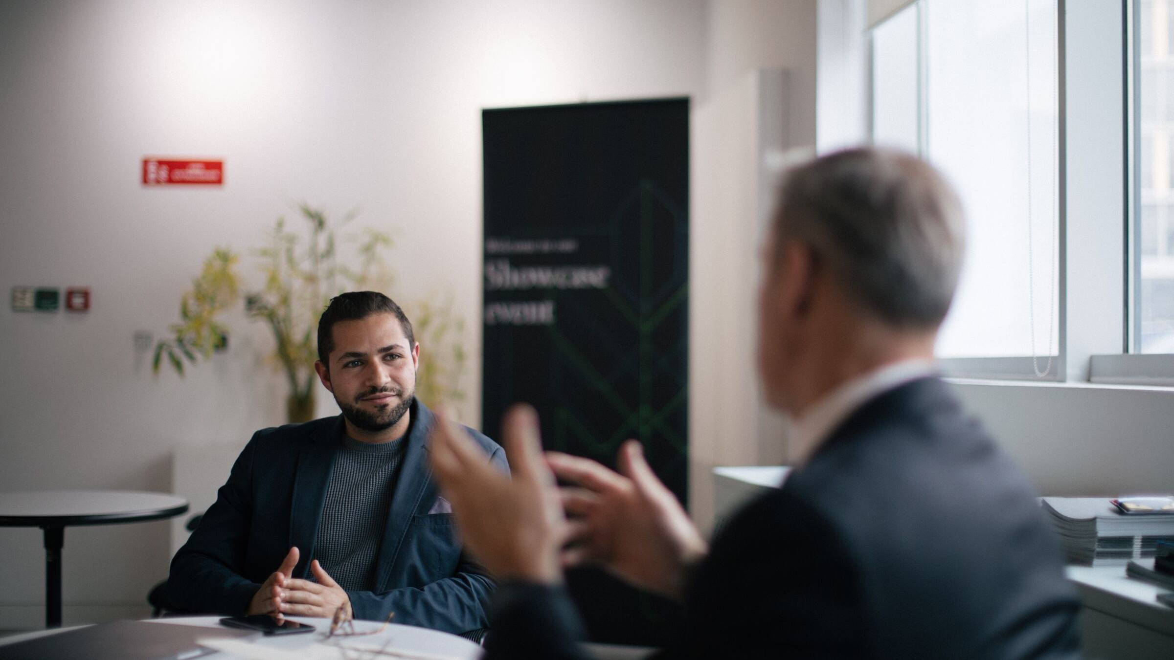 A man in a blazer seated at a table listening to a colleague in a suit who gestures as he speaks, at a business event