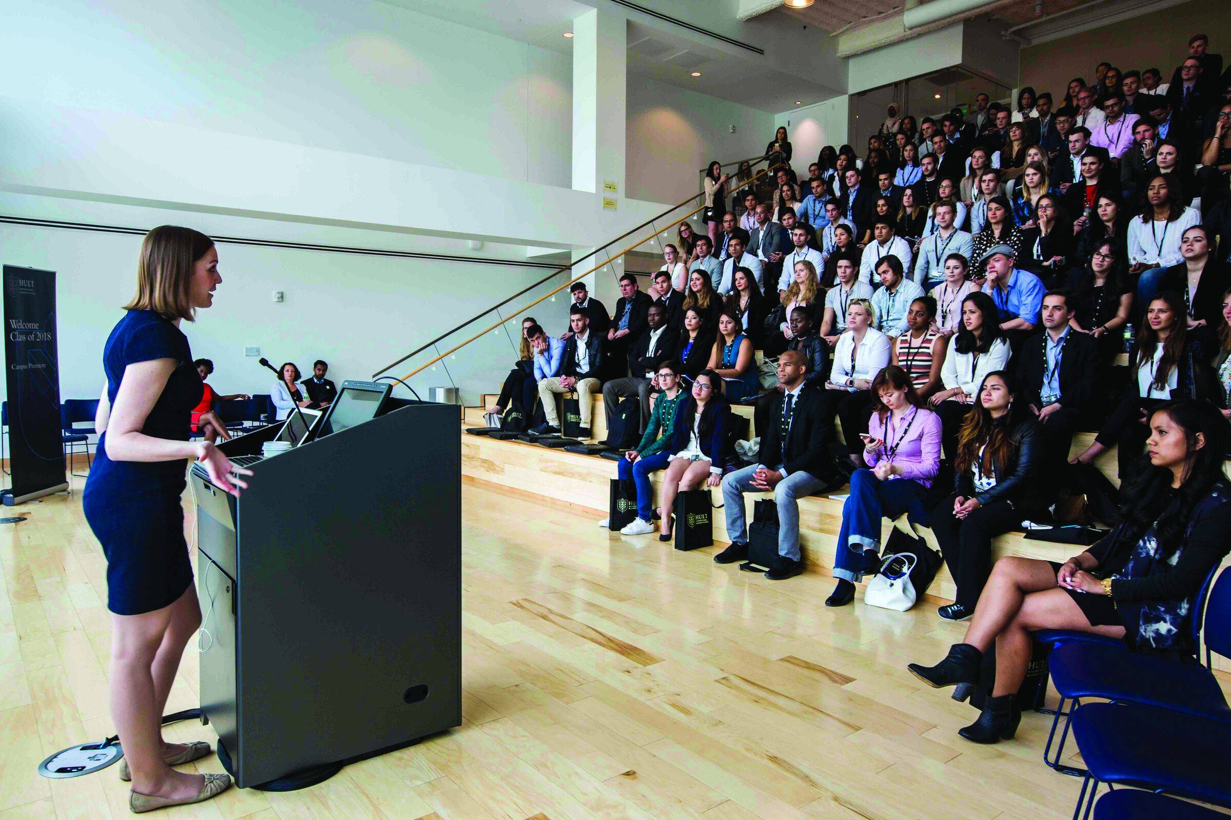 Woman speaking at a podium to a large seated audience in a bright auditorium