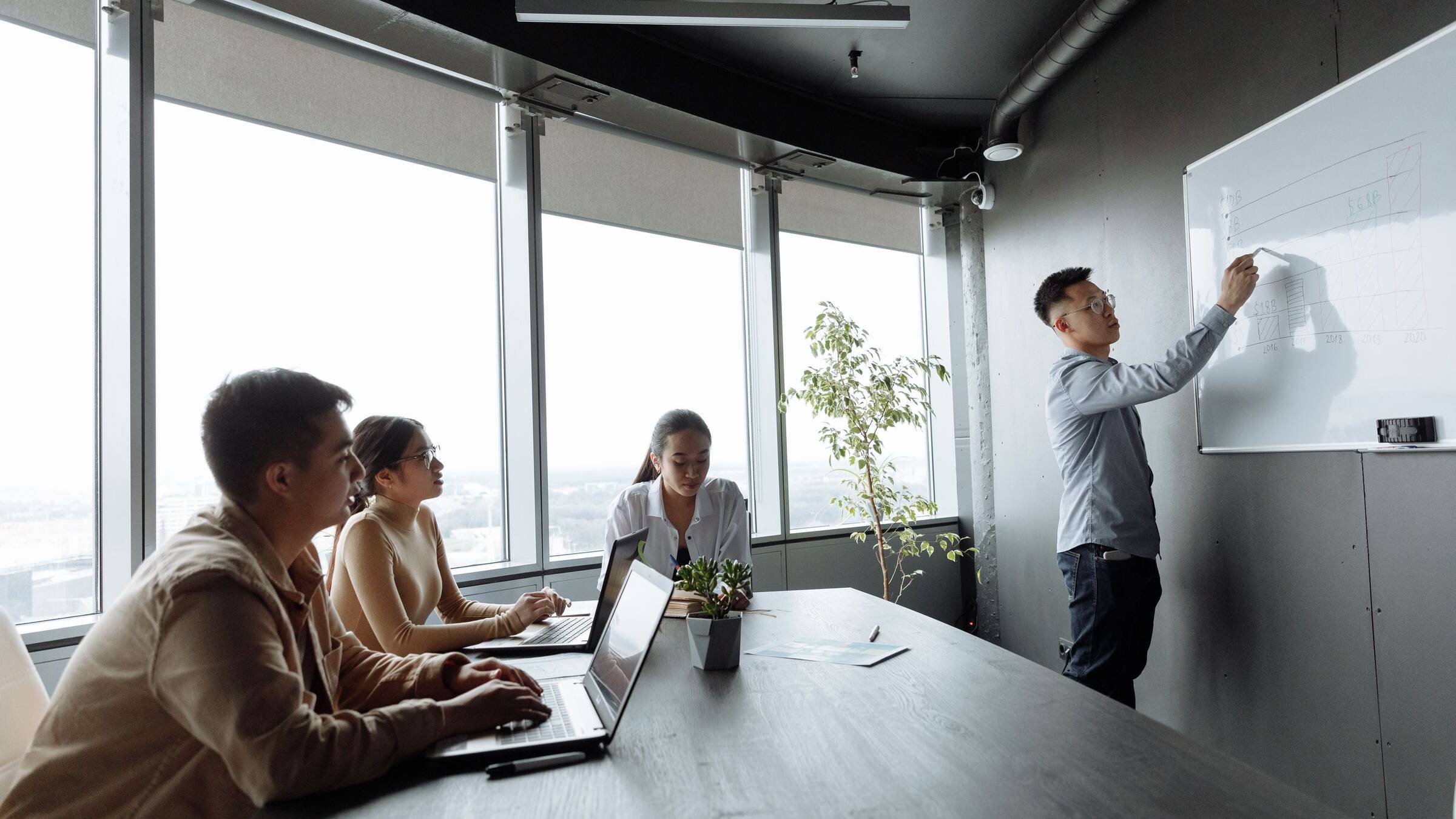 Man writing on a whiteboard in a glass-walled meeting room while three colleagues at a table with laptops look on
