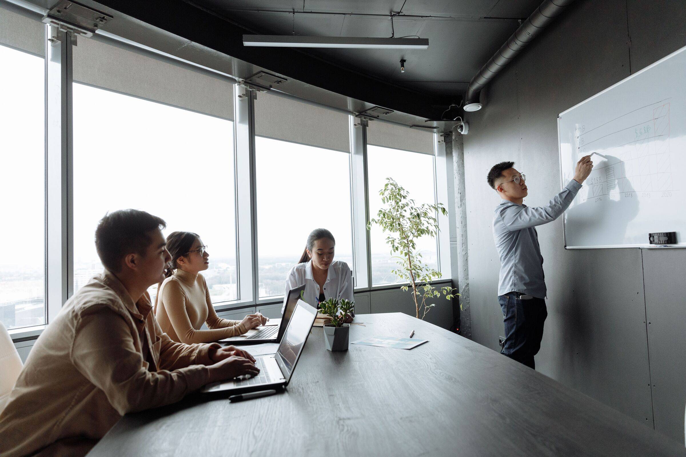 Man writing on a whiteboard in a glass-walled meeting room while three colleagues at a table with laptops look on