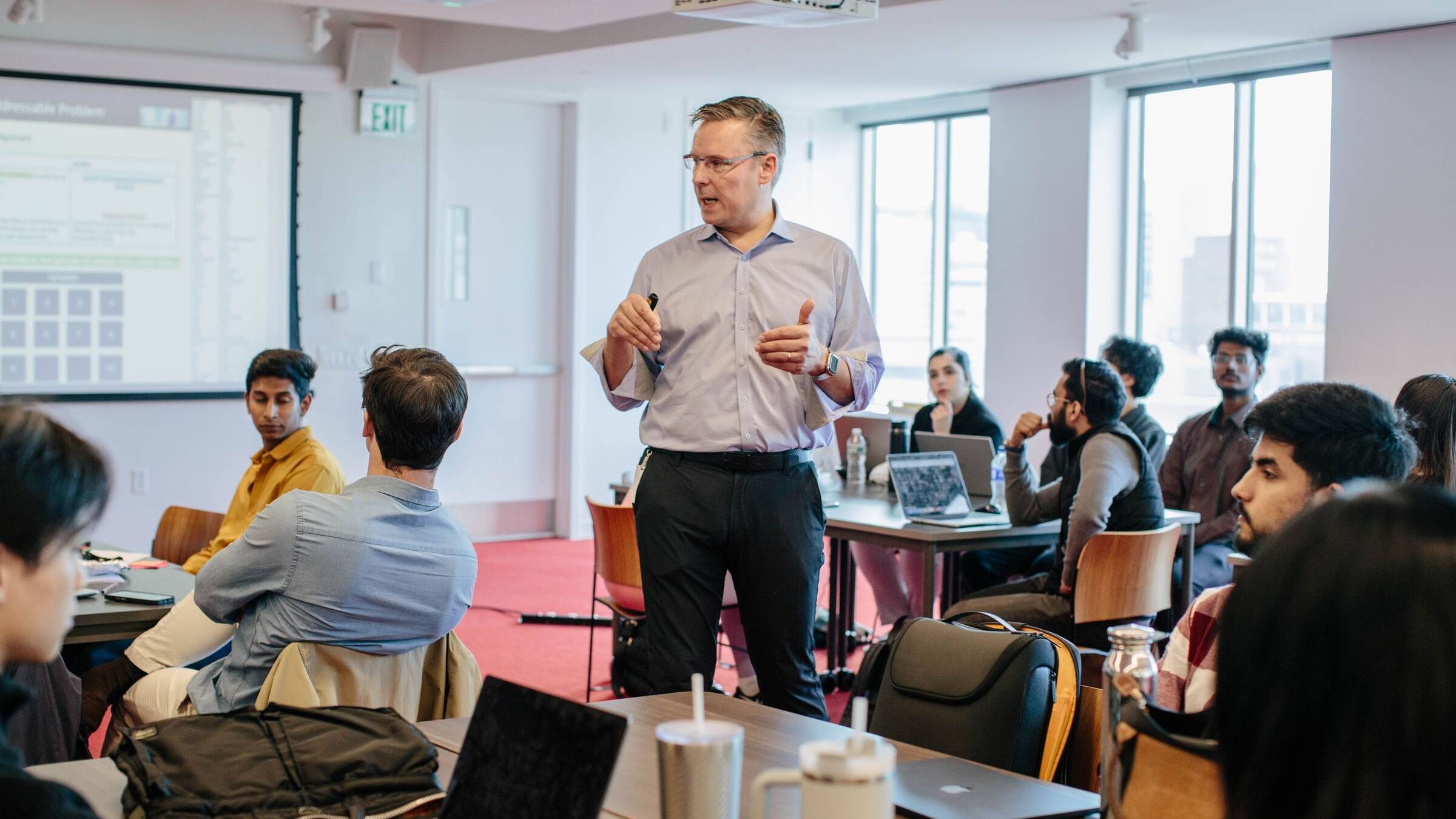 Lecturer standing in a bright seminar room, speaking to students seated at tables with laptops