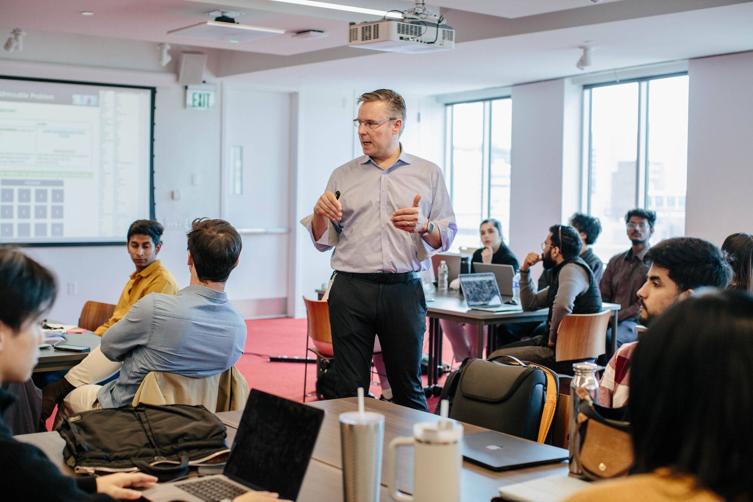 Lecturer standing in a bright seminar room, speaking to students seated at tables with laptops