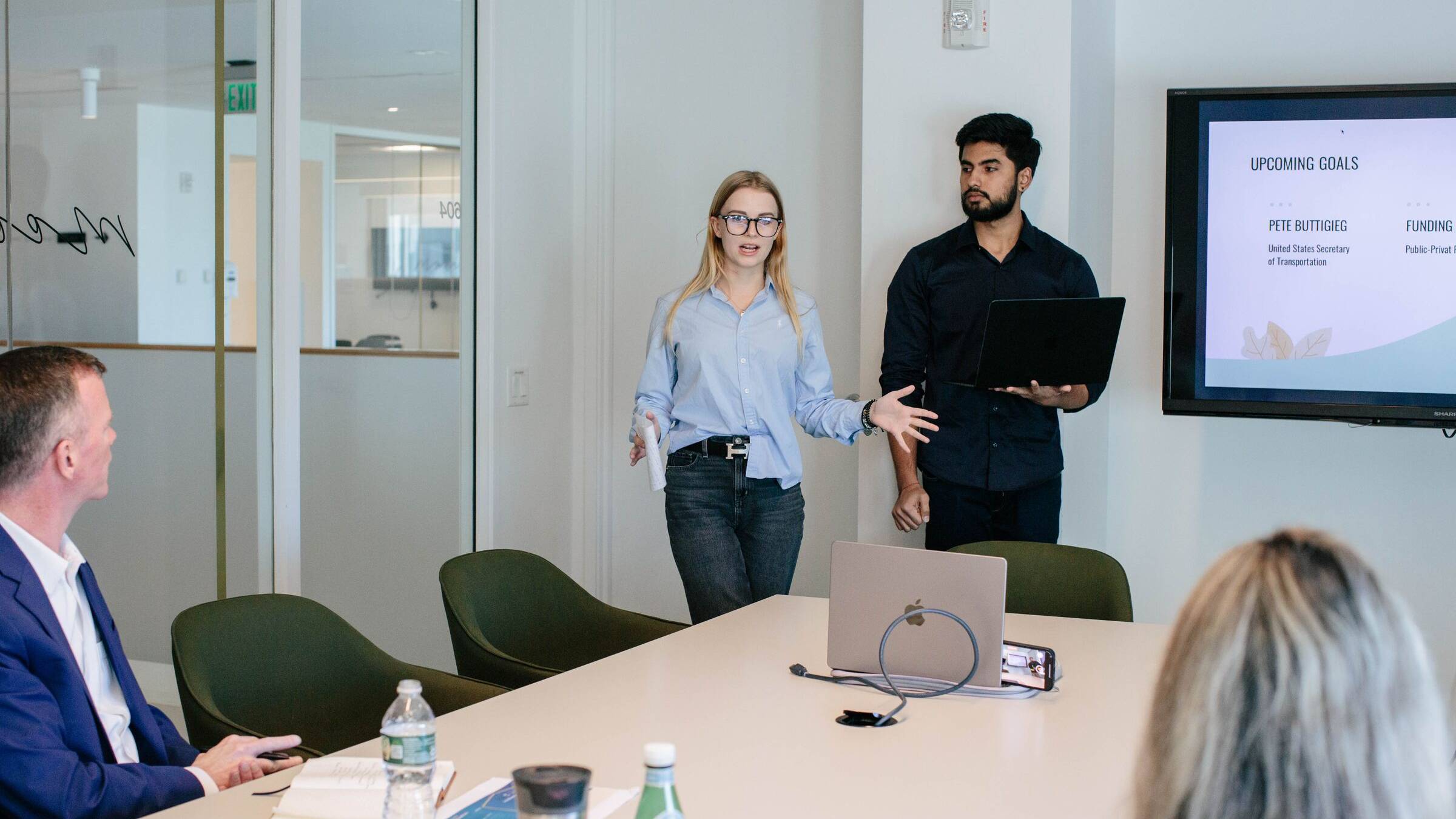 Two students presenting to a small group seated around a boardroom table, with a slide on the screen behind