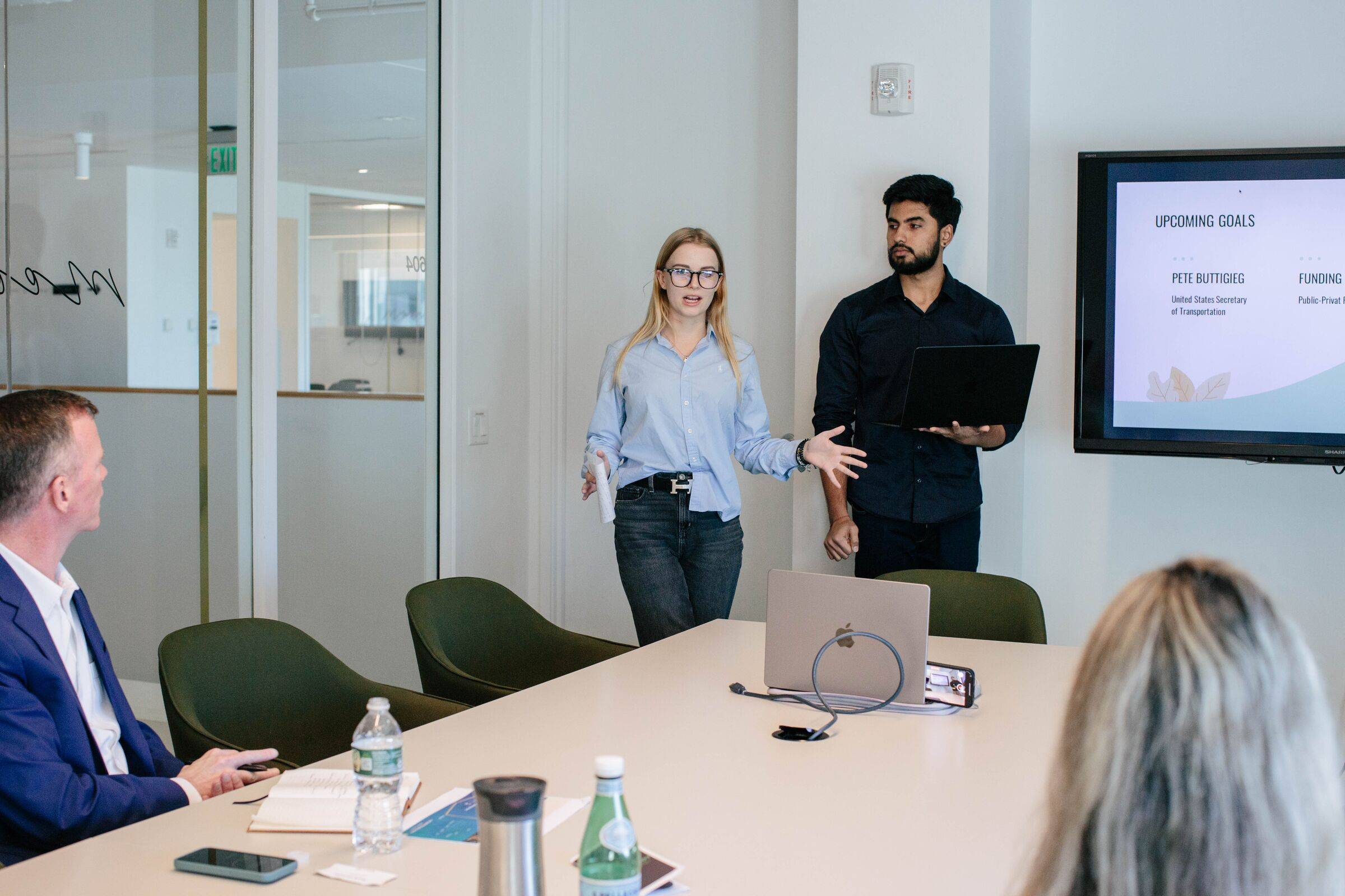Two students presenting to a small group seated around a boardroom table, with a slide on the screen behind