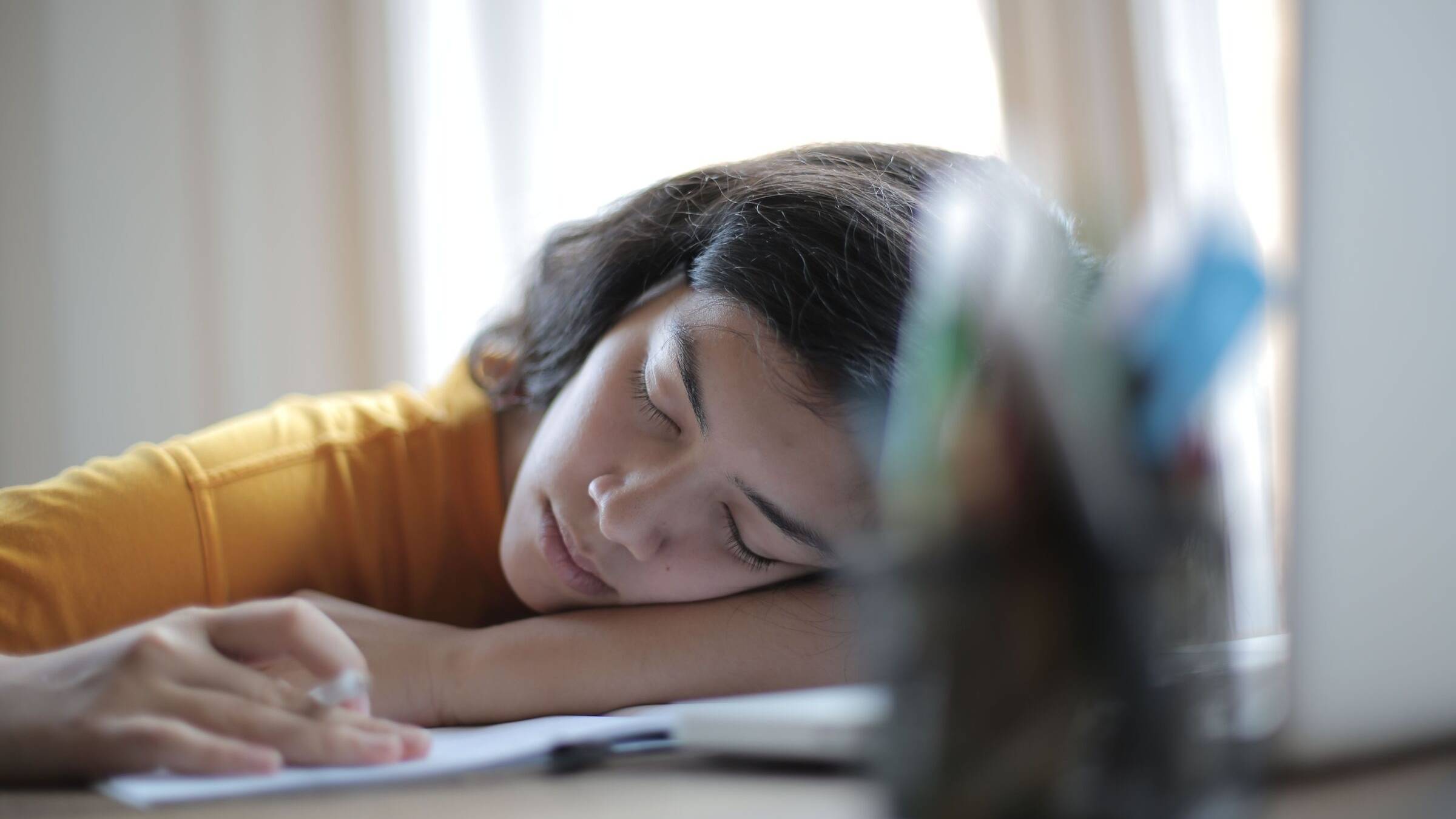 Student asleep with her head resting on her arm at a desk, a notebook and pen in front of her