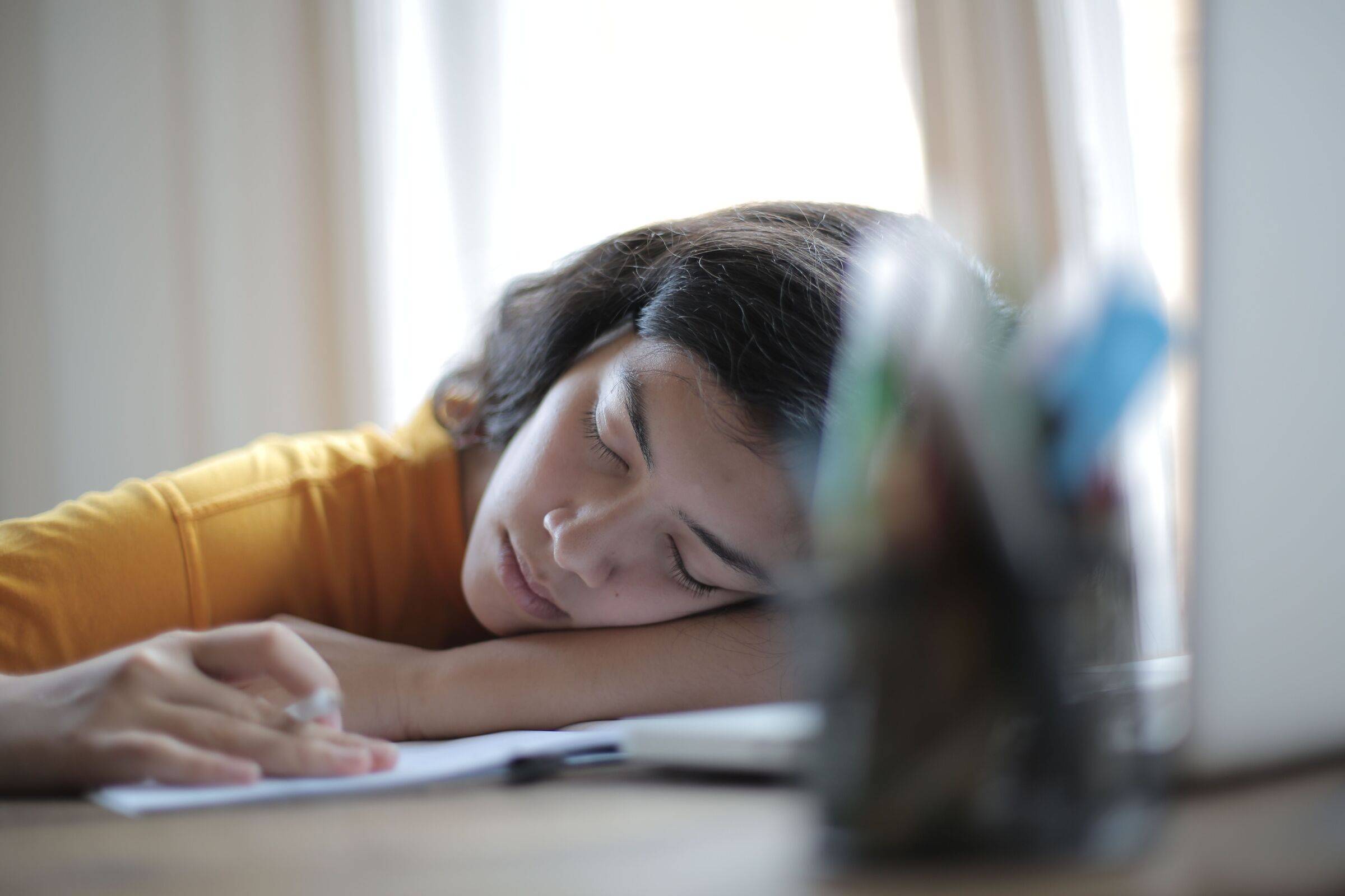 Student asleep with her head resting on her arm at a desk, a notebook and pen in front of her