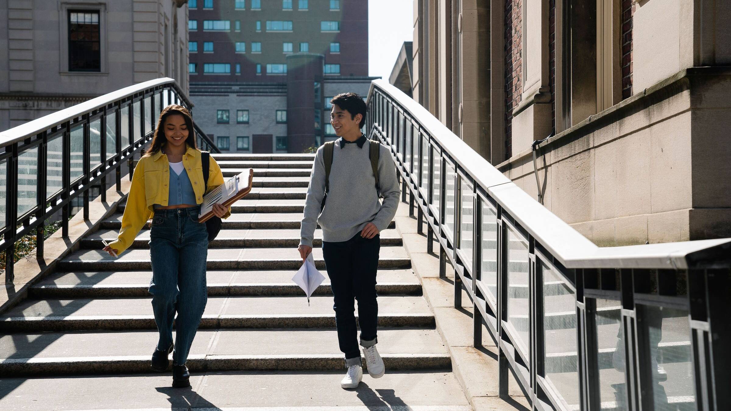 Two students walking down a flight of campus steps carrying books and bags