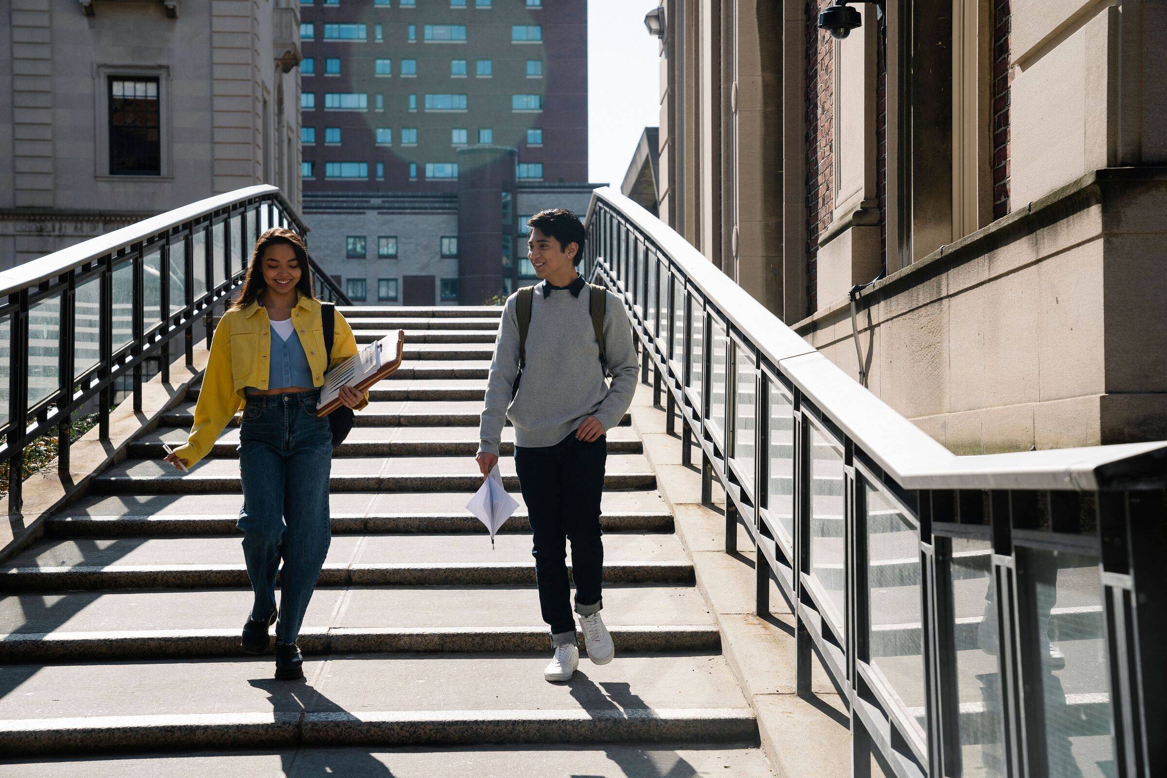 Two students walking down a flight of campus steps carrying books and bags