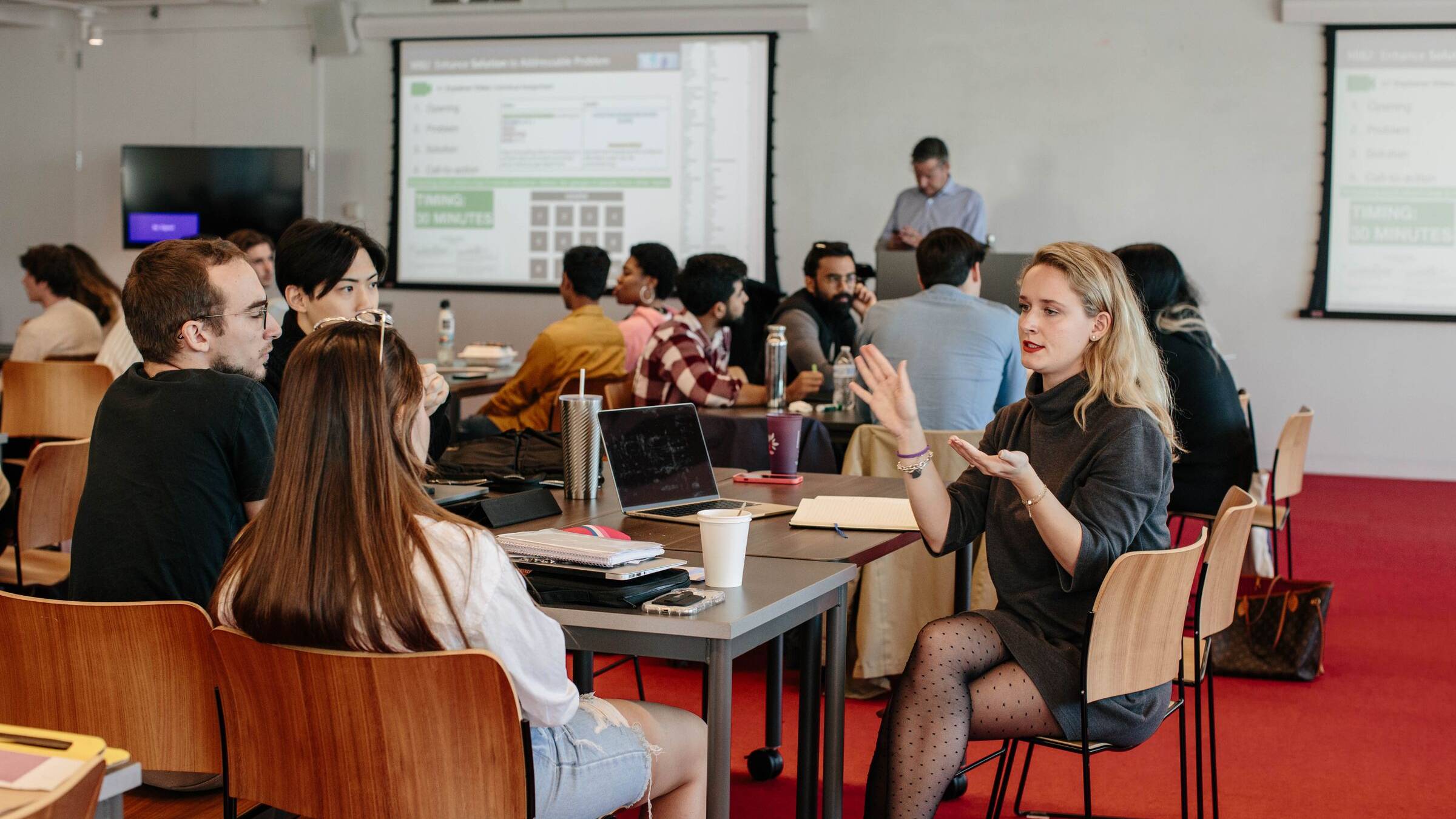 Students in a seminar room discussing around tables while a presenter stands at the back beside a screen showing data