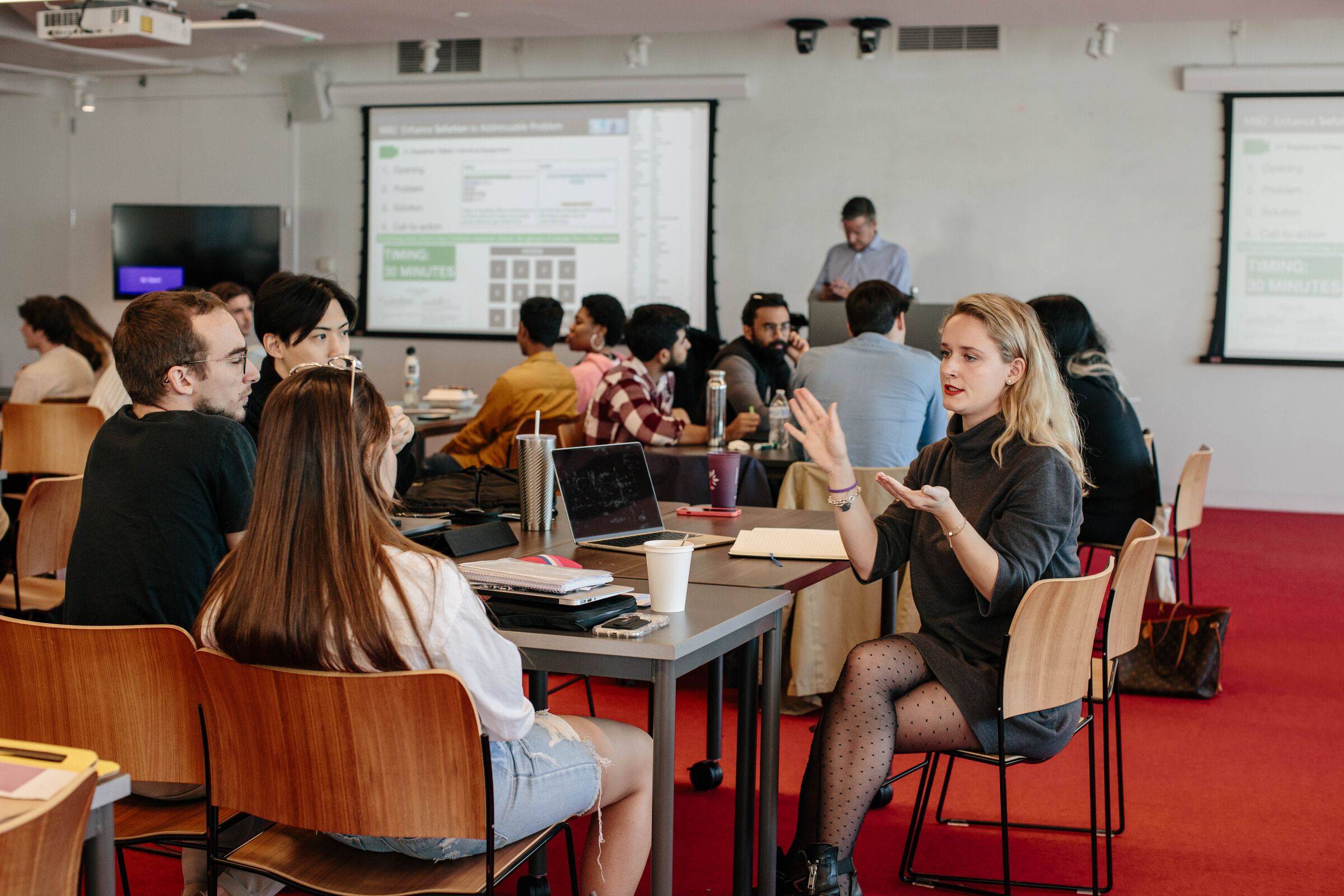 Students in a seminar room discussing around tables while a presenter stands at the back beside a screen showing data