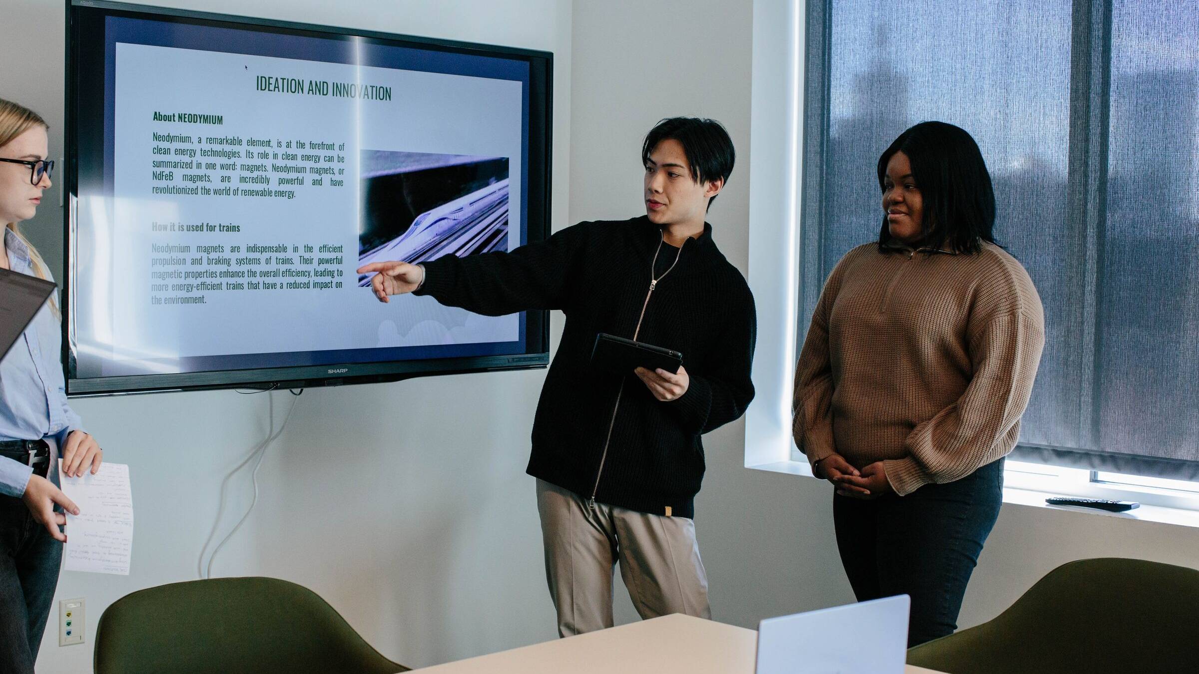 Two students presenting a slide on a wall-mounted screen in a meeting room
