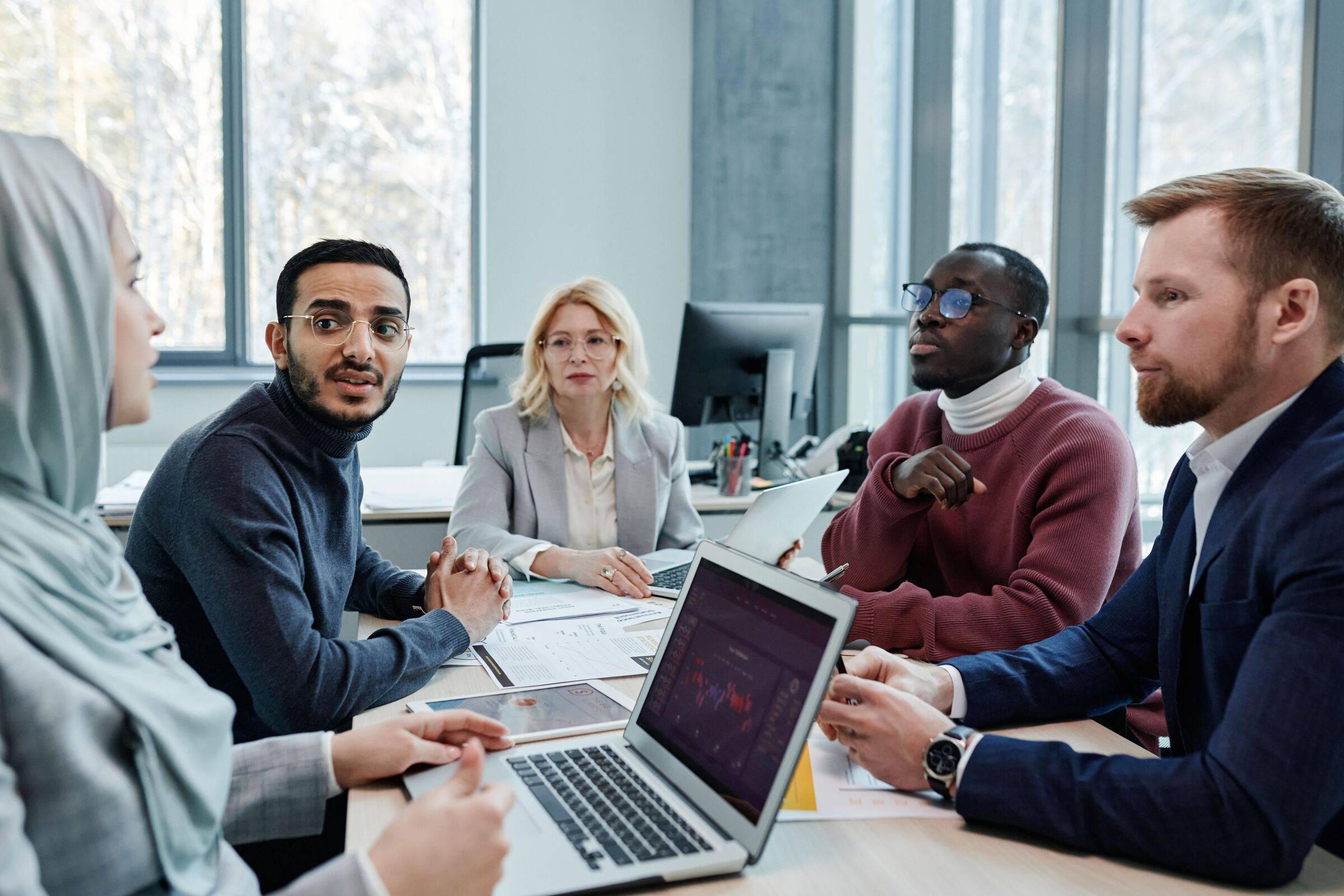 Five colleagues from different backgrounds in discussion around a meeting table with laptops and papers