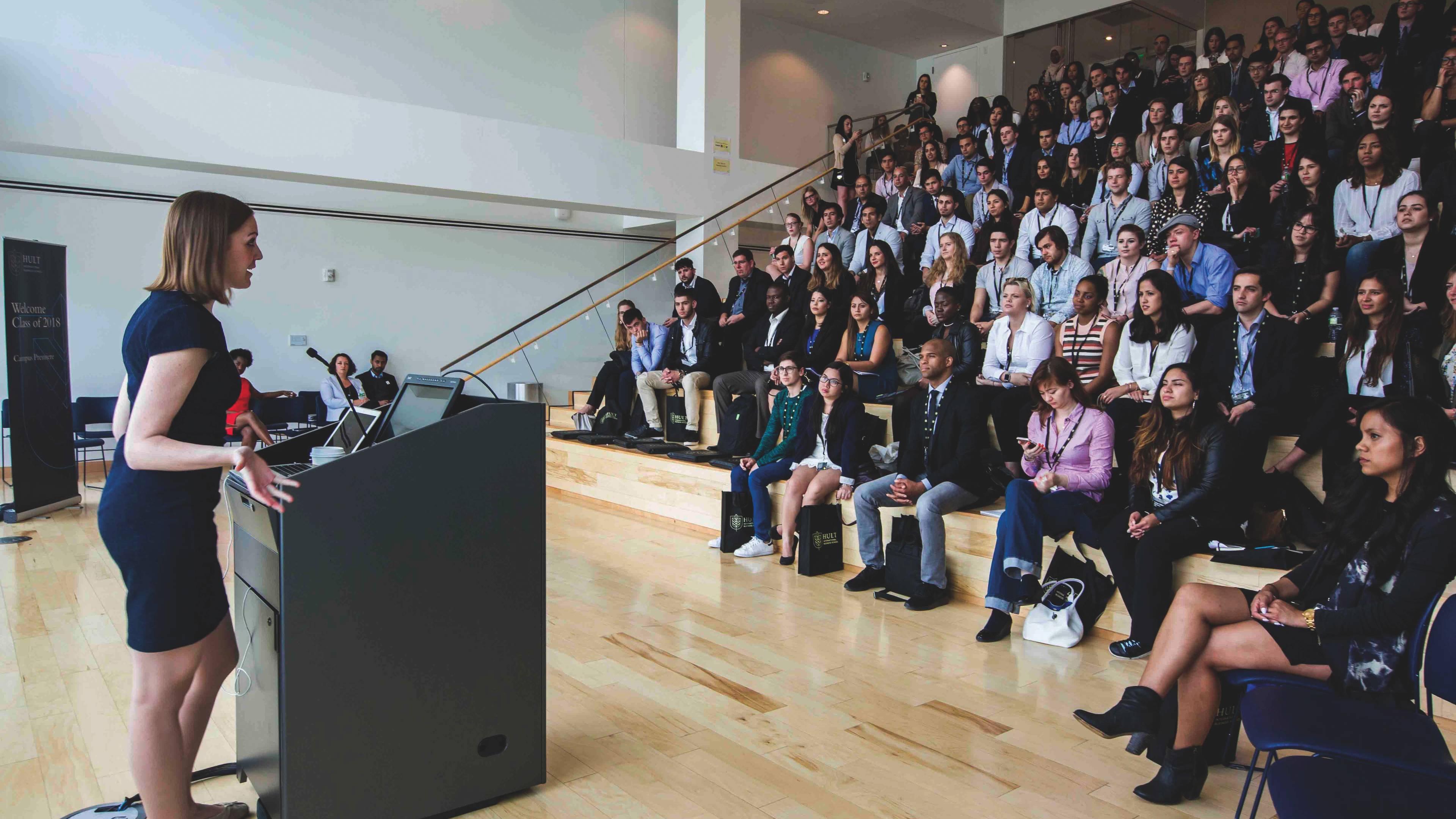 A woman in a blue dress speaks at a podium to a seated audience in a lecture hall with tiered seating.