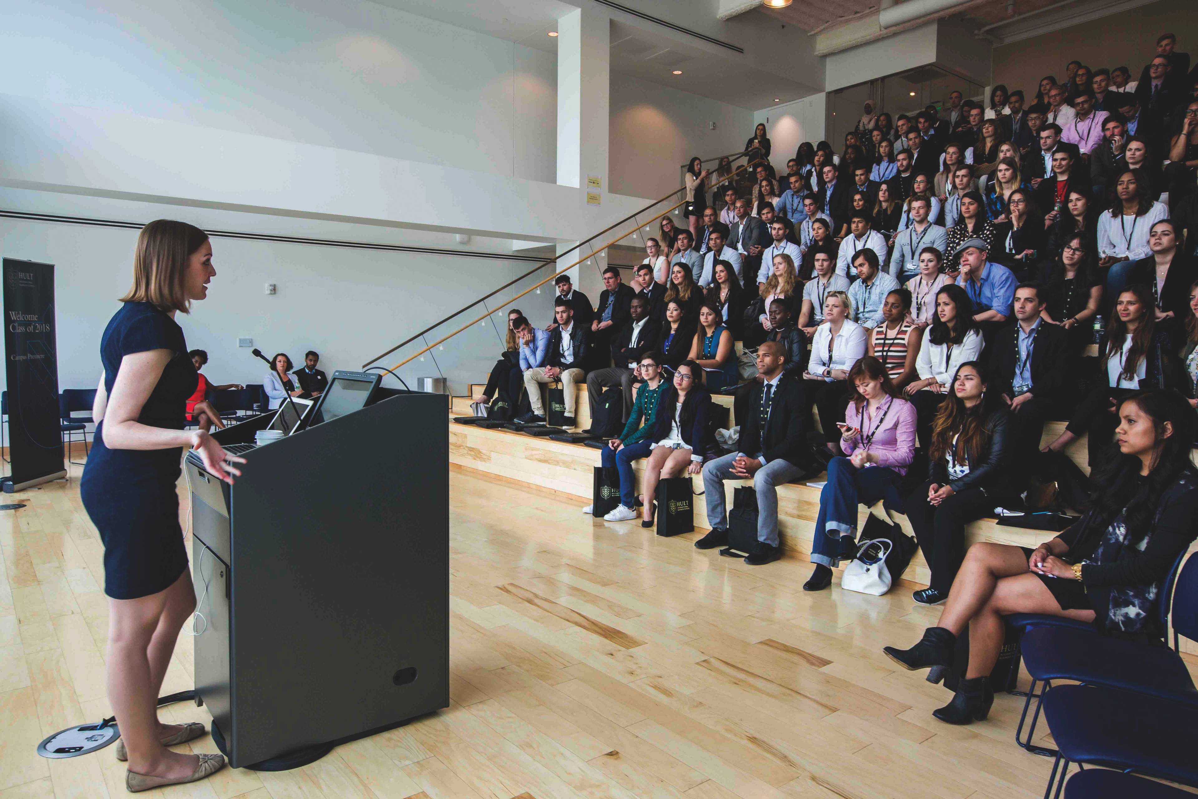 A woman in a blue dress speaks at a podium to a seated audience in a lecture hall with tiered seating.