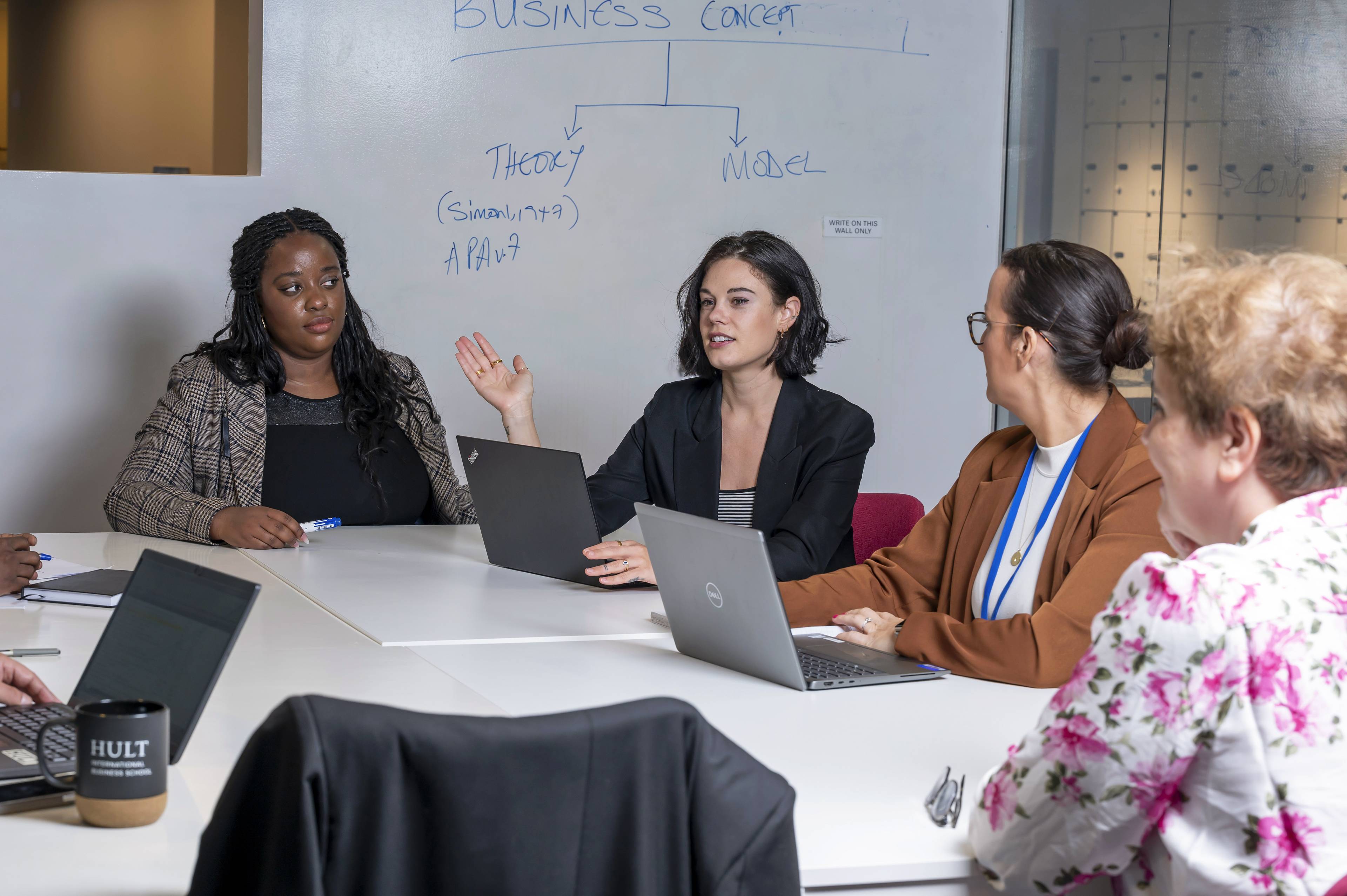 A group of diverse people in a meeting room, discussing with laptops open. One person gestures while speaking, with a whiteboard behind them.