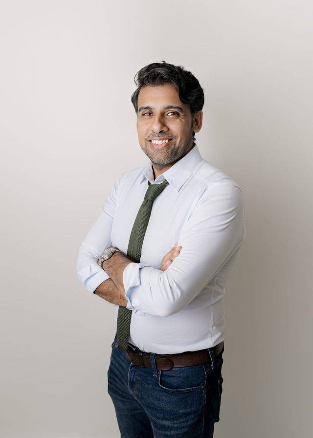 A smiling man with crossed arms, wearing a white shirt, green tie, and jeans, stands against a plain background.