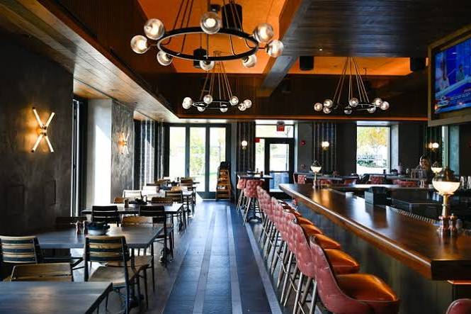 Empty modern restaurant interior with pendant lights, wooden tables, and a bar with red stools. Large windows and a TV screen are visible.