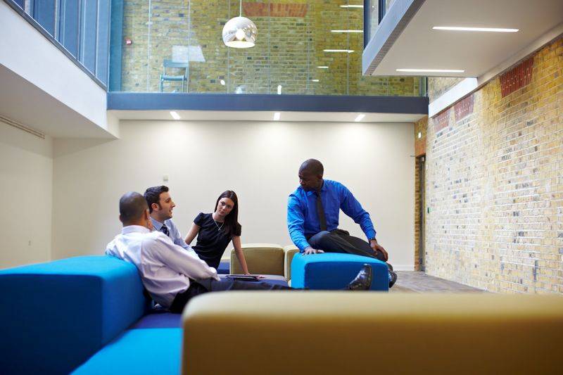 Four people having a casual meeting on colorful couches in a modern campus with brick walls and large windows.
