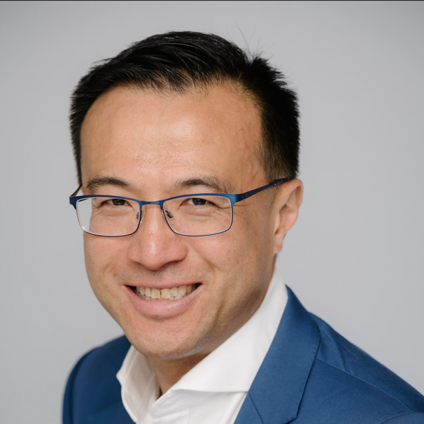 Smiling man in glasses and a blue suit against a gray background.
