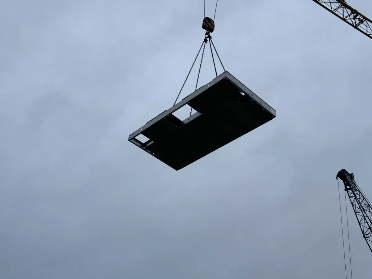 Een pre-made zoldervloer hangt aan de kraan boven de bouwplaats, met de sparing er al in.