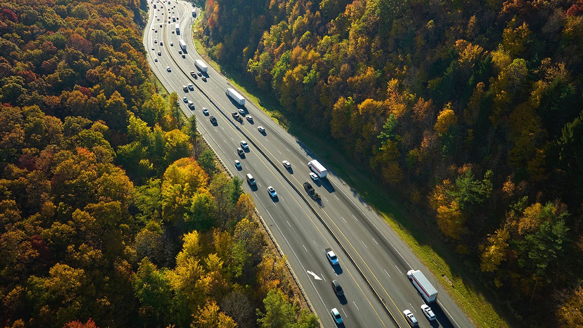 Aerial view of a highway with cars and trucks, surrounded by dense autumn-colored forest.