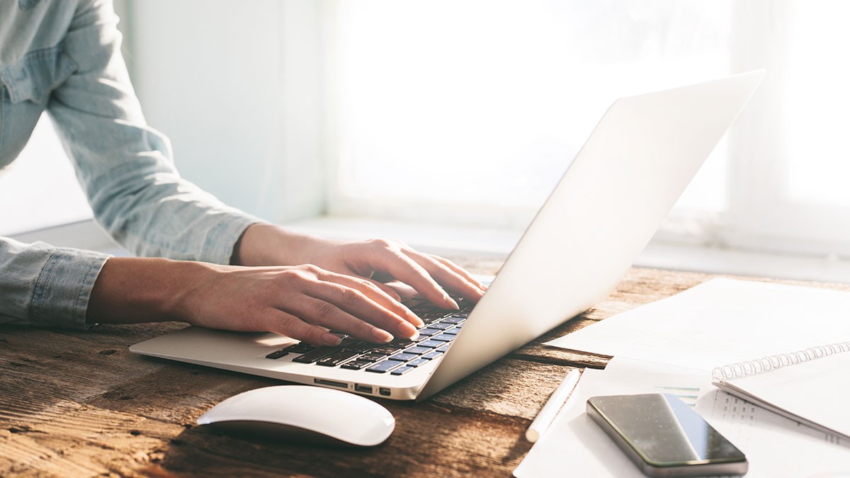 Person typing on a laptop at a sunlit wooden desk, with a mouse, smartphone, and notebooks nearby.