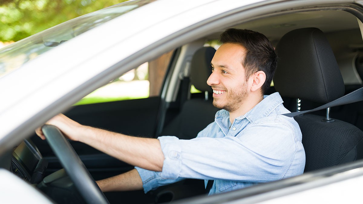 A smiling man in a light blue shirt is driving a car, looking forward through the windshield.