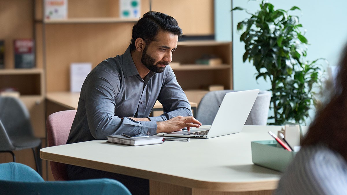 A man sits at a desk with a laptop and a stack of books, focusing intently on his work in a modern office setting with a plant nearby.