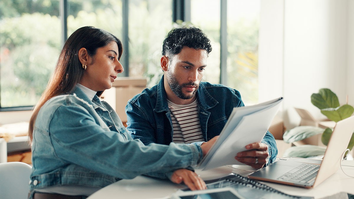 Two people sitting at a table, reviewing documents together, with a laptop open and a bright window in the background.