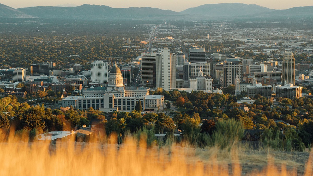 Aerial view of a cityscape with a prominent capitol building surrounded by skyscrapers, set against a backdrop of mountains and a golden field.